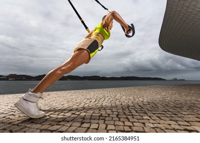 Woman doing push ups with trx fitness straps outdoors. Sporty woman with fit body in sportswear posing on sky background.
