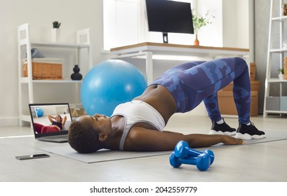 Woman Doing Sports Workout At Home. Black Lady Watching Video Lesson, Having Virtual Class With Remote Online Instructor On Notebook PC And Doing Strengthening Glute Bridge Gymnastics On Exercise Mat