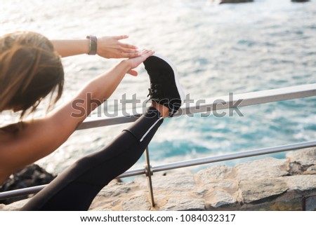 A woman doing stretches by the sea side touching her toes and wearing black tights and sneakers