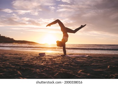 Woman doing yoga pose handstand with splits on the beach with sunset in background. Female exercising yoga on the sea shore.