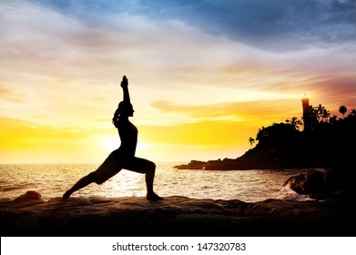 Woman Doing Yoga Warrior Pose In Silhouette On The Cliff Near Lighthouse At Sunset Sky In Kovalam, Kerala, India