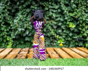 Woman doll with dark skin holding a photo camera in front of her face, talking a picture. In the garden, blurred background. 