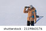 Woman in down jacket with a backpack and ski poles in hand looks through binoculars standing on a snowy plain in winter, selected focus.