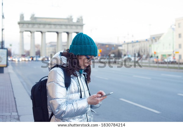 woman\
in a down jacket and hat stands at a public transport stop and uses\
a smartphone. Wait for the bus. Choose a route in the mobile app,\
search for a bus route or call a taxi on your\
phone