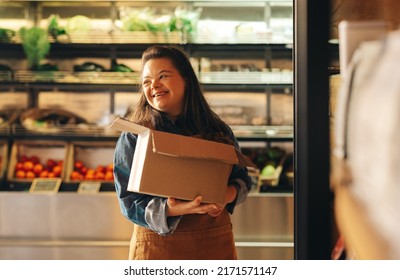 Woman with Down syndrome smiling happily while working as a shopkeeper in a grocery store. Empowered woman with an intellectual disability restocking food products in a supermarket. - Powered by Shutterstock