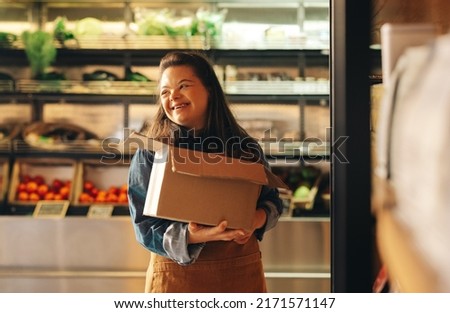 Woman with Down syndrome smiling happily while working as a shopkeeper in a grocery store. Empowered woman with an intellectual disability restocking food products in a supermarket.