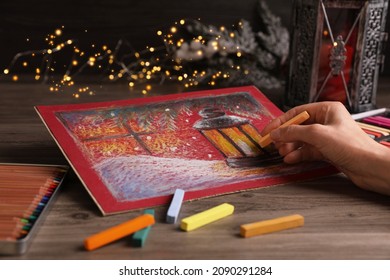 Woman drawing beautiful lantern with soft pastel at wooden table against blurred lights, closeup