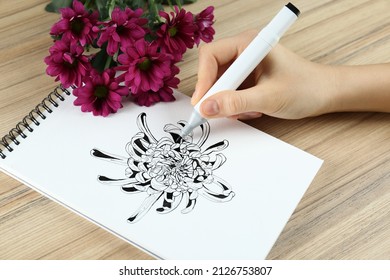 Woman drawing chrysanthemum in sketchbook and flowers at wooden table, closeup