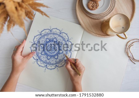 A woman draws a doodle mandala on a white sheet. Relax concept. White wooden table. Aged wood. A mug of coffee and chocolate chip cookies. Rosary and wooden beads.