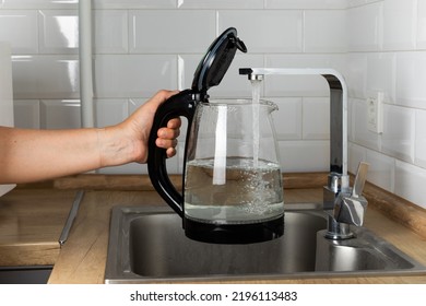 A woman draws water into an electric kettle in the background of the kitchen. It's time for breakfast and tea. Modern electric kettle on a wooden table. Kettle for boiling water.