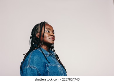 Woman with dreadlocks looking away thoughtfully while standing against a grey background. Stylish mature woman wearing a denim jacket and make-up in a studio. - Powered by Shutterstock