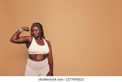Woman with dreadlocks looking at the camera while flexing her bicep. Mature woman standing against a brown background in sports clothing. Sporty black woman maintaining a fit lifestyle. - Shutterstock ID 2177474313