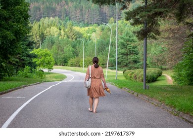 A Woman In A Dress With A Handbag On Her Shoulder And A Braided Ponytail Walking Down An Empty Road Barefoot Carrying Her Shoes In Her Hands