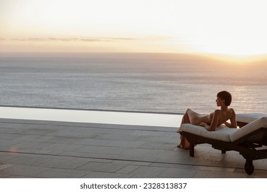 Woman in dress laying on lounge chair on patio overlooking ocean at sunset - Powered by Shutterstock