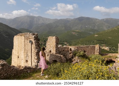 Woman in dress looking at ruins of an ancient stone building in Himare Castle, Vlore, Albania. Meadow of wildflowers blooming with scenic view of Ceraunian mountain range. Collapsed historical site