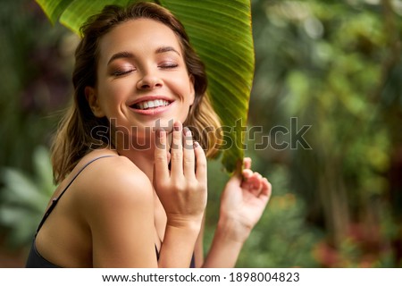 woman in dress shine with happiness, enjoy being in forest touching face, she is smiling, having perfect skin and toothy smile. around green plants