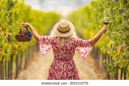 Woman in dress and straw hat has rised hands and holds a glass of delicious red wine and a basket full of grapes. She walks through a vineyard. - Powered by Shutterstock