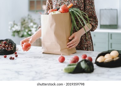 Woman in dress unpacking vegetables from paper grocery bag onto kitchen island with marble top, getting ready to prepare healthy and nutritious meal, furniture in blurred background, cropped shot