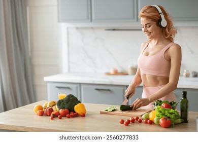 A woman dressed in athletic clothing is chopping vegetables in a contemporary kitchen. She enjoys music through headphones while surrounded by fresh ingredients like cucumbers and tomatoes, copy space - Powered by Shutterstock