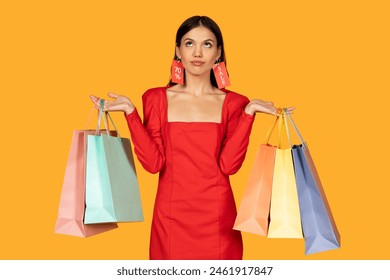 A woman dressed in a chic red dress stands against a warm yellow background, her hands holding several shopping bags in various pastel shades. Bright red sale tags are attached to her ears - Powered by Shutterstock