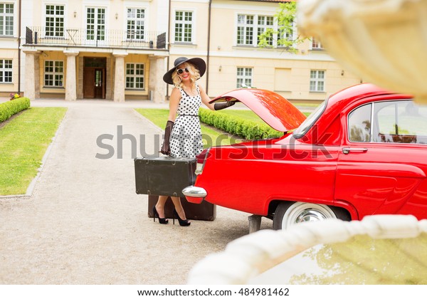 Woman dressed in cocktail dress holding vintage\
suitcase next to red retro\
car