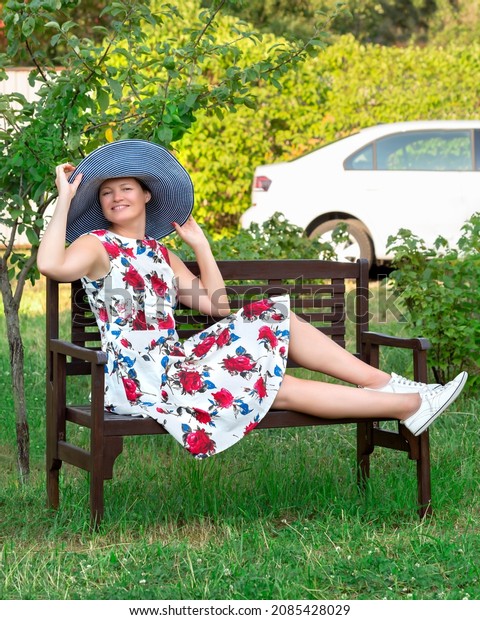 A woman dressed in a floral dress and hat is\
sitting on a bench in the\
garden