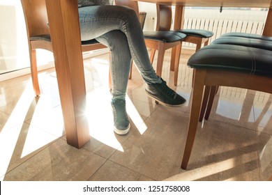 A Woman Dressed In Jeans. Woman's Legs Under The Table