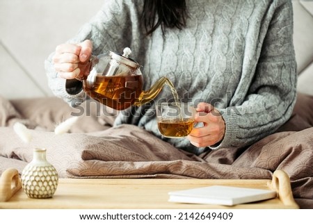 Woman dressed in knitted sweater pours hot tea from glass teapot into mug while sitting in bed. Morning breakfast in cozy home bedroom interior. Hygge, warm, autumn concept.