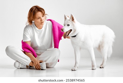 A woman dressed in white with a pink wrap sits on the floor next to her white Siberian Husky. The scene captures a moment of quiet companionship in a minimalist studio - Powered by Shutterstock