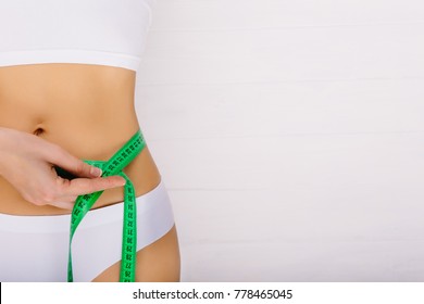 woman dressed in white underwear measures her slim stomach on a light wooden background with empty space. concept ideal body, sport training and diet.