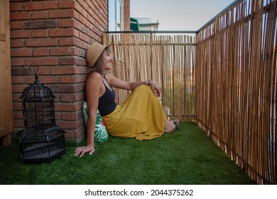 Woman dressed in a yellow skirt and black tank top sitting on the lawn of the terrace of her house looking at the horizon. Resting.