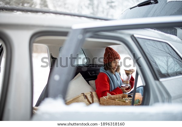 Woman drink coffee in car trunk, traveling\
by rent car during winter holidays. View from the window, from the\
outside of the car. High quality\
photo