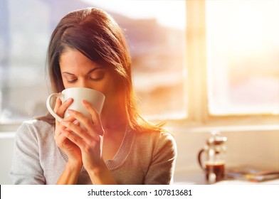 Woman drinking coffee at home with sunrise streaming in through window and creating flare into the lens. - Powered by Shutterstock