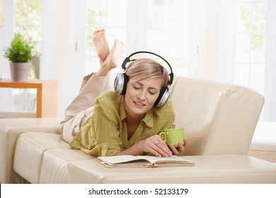 Woman Drinking Coffee, Lying On Couch Reading Book, Wearing Headphones At Home.