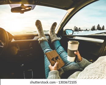 Woman drinking coffee paper cup inside car with feet warm socks on dashboard - Girl relaxing in auto trip reading travel book with snow mountains in background - Traveler concept - Focus on feet