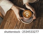 Woman drinking coffee sitting by the textured wooden table. Close up hands with cup of cappuccino.