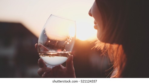 Woman Drinking Cooled White Wine from a glass at the Balcony during Sunset.  Beautiful female enjoying cozy evening on terrace. Summertime relax at sunny patio. Lens Flare