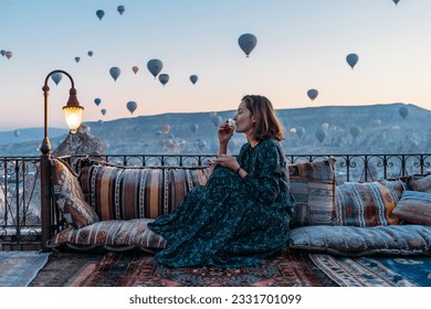 Стоковая фотография: Woman drinking early morning tea with hot air balloons in Cappadocia