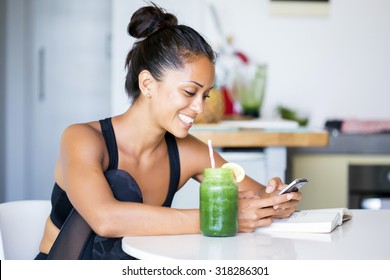 Woman drinking a homemade green detox juice, wearing sportive clothing, texting on her  phone while sitting in her kitchen table