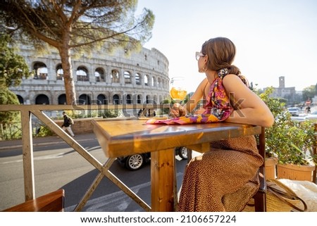 Woman drinking Spritz Aperol at outdoor cafe near Coliseum, the most famous landmark in Rome. Concept of italian lifestyle and traveling Italy. Caucasian woman wearing dress and shawl in hair