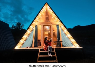 Woman drinking tea on the porch of a wooden lodge with lights of garlands in the evening. The concept of glamping and renting a chalet for weekend