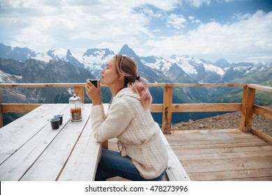 Woman drinking warm tea in the rustic wooden outdoor cafe on mountain, alpine view, snow on hills. Dombay, Karachay-Cherkessia, Caucasus, Russia. - Powered by Shutterstock