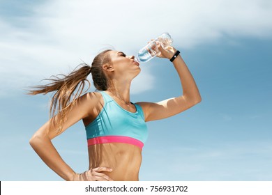 Woman Drinking Water After Running. Portrait Of Beautiful Athletic Girl In Bright Colorful Sportswear Resting After Fitness Workout, Drink Water From Bottle On Blue Sky Background. High Quality Image