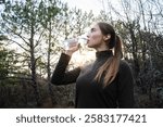 Woman drinking water from a plastic bottle standing in a forest. Hydration during sport activities, maintaining water balance. Stay hydrated. Wellness and healthy lifestyle outdoor. Low-angle portrait