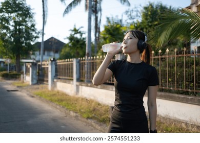 A woman is drinking water while walking down the street. She is wearing headphones. The street is lined with palm trees