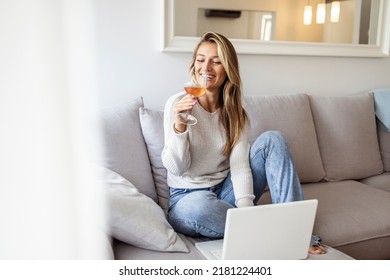Woman drinking wine while using laptop. Young female is sitting at sofa. She is wearing striped t-shirt at home. Brunette sitting on couch in living room, drinking wine and using laptop.