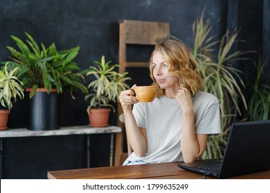 A Woman Drinks Coffee And Enjoys At Her Work Desk With A Laptop In A Cozy Home Office With Tropical Plants In The Background. .Working Freelancer.