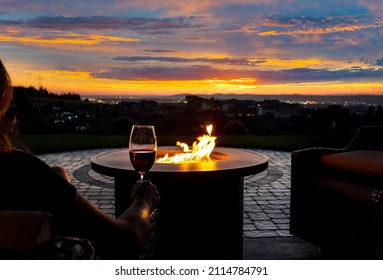 A woman drinks a glass of wine in front of a fire pit on the luxury patio of a hillside home overlooking city lights.	 - Shutterstock ID 2114784791