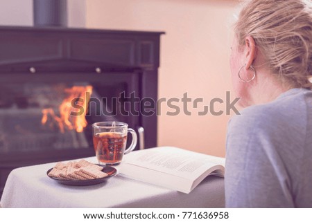 A woman drinks tea and reads a book in front of the fireplace