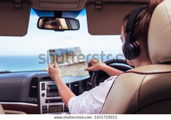 Woman driver in the\
headphones driving a car and looking at th map. Girl relaxing in\
auto trip traveling along ocean tropical beach in background.\
Traveler concept. 